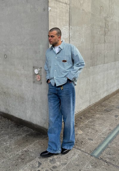 Camisa de botones a rayas en azul y blanco, combinada con jeans de pierna ancha de un azul claro y botas negras. De pie contra una pared de hormigón.