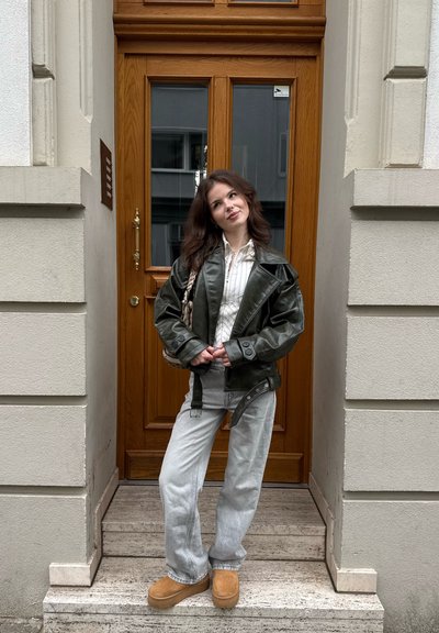 Chaqueta de cuero verde oscuro sobre una camisa blanca con rayas finas, combinada con pantalones vaqueros claros y zapatos de plataforma beige, de pie sobre escalones de piedra.