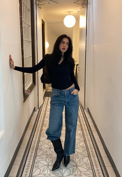 Top de manga larga oscuro, jeans de pierna ancha de color azul claro y botas negras hasta la rodilla. De pie en un pasillo estrecho con baldosas de suelo estampadas.