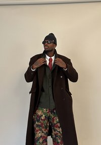 Muž v tmavě hnědém dlouhém kabátu, zelené zapnuté bundě, bílé košili s červenou kravatou, maskáčových kalhotách, černé pletené čepici a slunečních brýlích si upravuje límec kabátu.