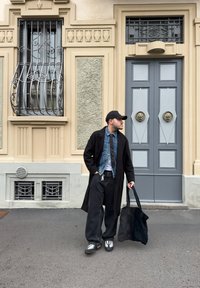 Osoba v dlouhém černém kabátu přes denimovou bundu, spárovaná s černými kalhotami a teniskami, stojící blízko šedých dveří s dekorativními akcenty.