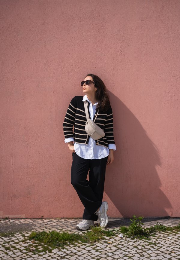 Striped black and cream cardigan over a white shirt, black pants, and light gray sneakers. Against a pink wall with a shadow cast.