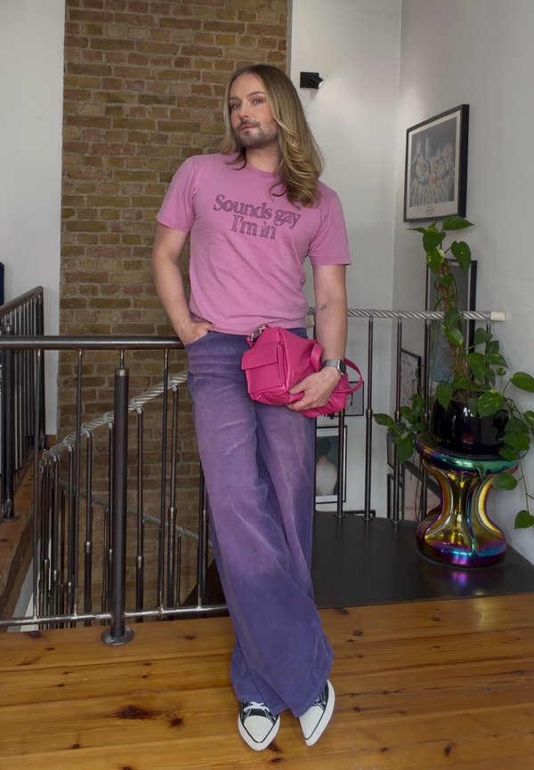 Personne avec de longs cheveux portant un t-shirt rose avec un texte, pantalon violet, chaussures blanches, tenant un sac rose, debout à l'intérieur près d'une rampe et de plantes.
