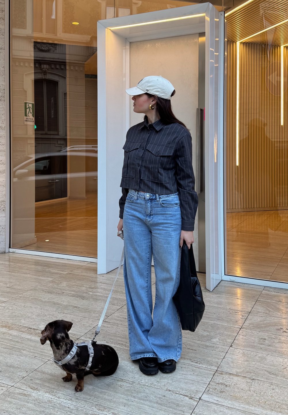 Camicetta nera a righe con due tasche, jeans a vita alta blu chiaro, scarpe con plateau nere e un cappello bianco. Un piccolo cane è al guinzaglio al suo fianco.