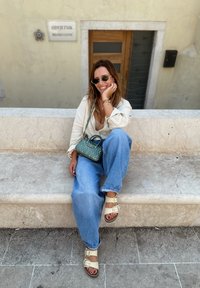 Jeans a gamba larga blu, camicia di lino bianca, sandali beige con fibbie e una piccola borsa verde. Persona seduta su una panchina di pietra.