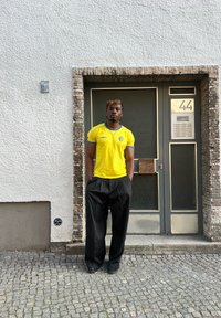 Κίτρινο κοντομάνικο πουκάμισο με μπλε λεπτομέρειες, μαύρο παντελόνι με ρίγες, στέκεται μπροστά σε έναν υφασμάτινο λευκό τοίχο και μια πόρτα με μια πινακίδα.