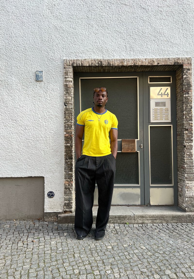 Κίτρινο κοντομάνικο πουκάμισο με μπλε λεπτομέρειες, μαύρο παντελόνι με ρίγες, στέκεται μπροστά σε έναν υφασμάτινο λευκό τοίχο και μια πόρτα με μια πινακίδα.