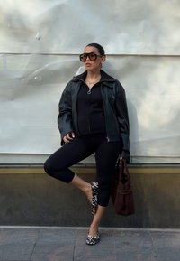 Žena opřená o zeď, oblečená v černé zapínací bundě, černých legínách, leopardích balerínách, velkých slunečních brýlích a držící hnědou kabelku.
