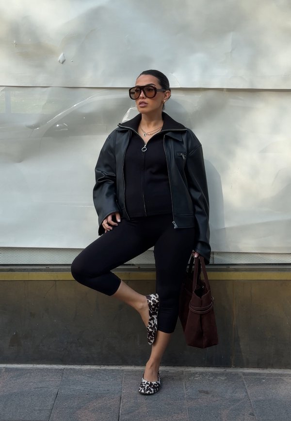 Nő, aki a falnak dől, fekete zipzáras dzsekit, fekete leggingset, leopárdmintás balerina cipőt visel, nagy napszemüveget hord és egy barna kézitáskát tart a kezében.