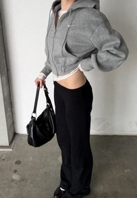 Sweat à capuche gris court avec zip et un logo blanc, superposé à un t-shirt léger à manches longues. Pantalon noir ample et sac à main noir.