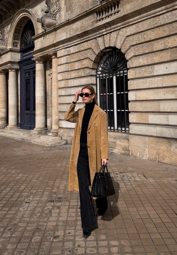 Femme portant un manteau beige, un col roulé noir et des lunettes de soleil, tenant un sac à main noir, marchant sur une rue pavée devant un bâtiment en pierre avec des fenêtres en arc.
