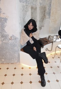 Mujer con cabello negro y botas sentada en un banco curvado, vestida con un conjunto negro y un chal de piel beige, en un café con suelo de baldosas estampadas.