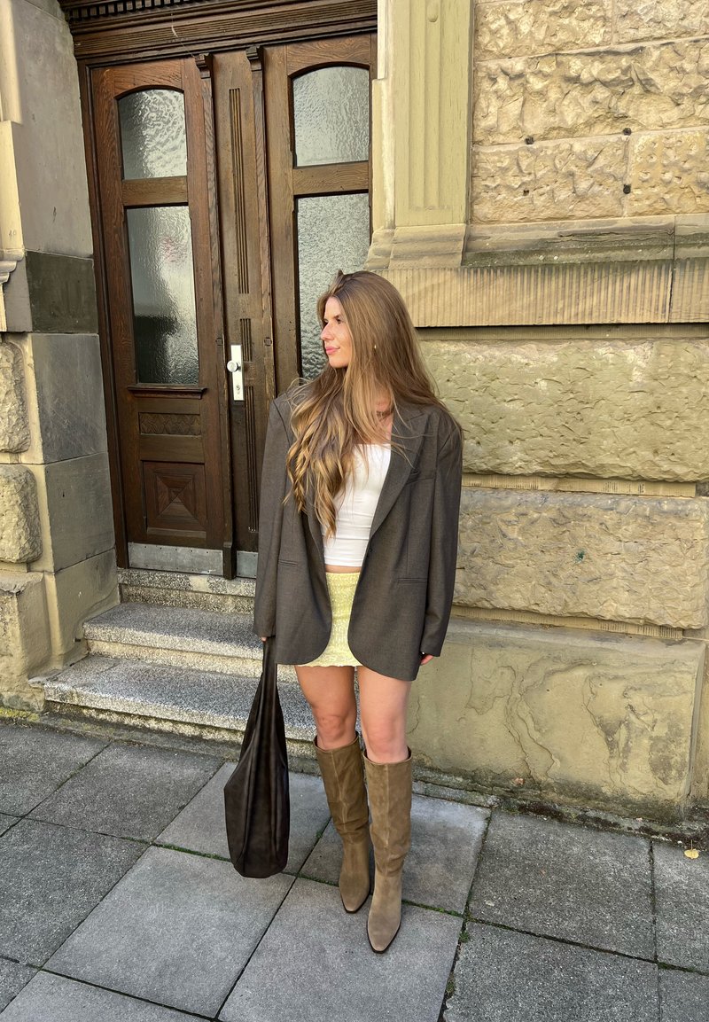 Jeune femme aux longs cheveux portant un blazer, un haut blanc, une jupe jaune et des bottes montant jusqu'aux genoux, tenant un sac noir, debout près de l'entrée d'un bâtiment en pierre.
