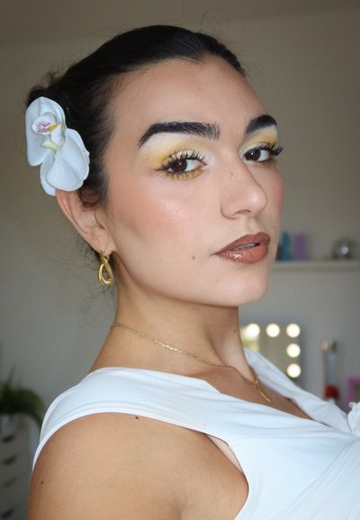 La modelo lleva una blusa sin mangas de color blanco. Presenta un degradado de sombra de ojos amarilla, labios marrones brillantes y un accesorio para el cabello en forma de orquídea blanca. También lleva pendientes de aro dorados.