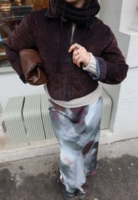 Άτομο που φοράει σκούρο καφέ μπουφάν, ανοιχτό πλεκτό πουλόβερ, πολύχρωμη σατέν φούστα, κρατώντας καφέ τσαντάκι, που στέκεται σε πεζοδρόμιο.