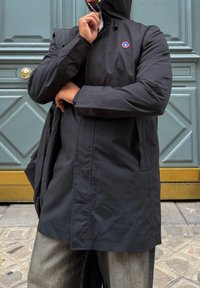 Black waterproof jacket with a hood, featuring a round red and blue logo on the chest, worn over grey denim trousers, against a textured wall.
