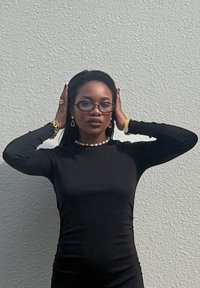 Black long-sleeve top, smooth texture, fitted shape. Accessories include a pearl necklace, gold earrings, bracelet, and ring. Clear glasses worn.
