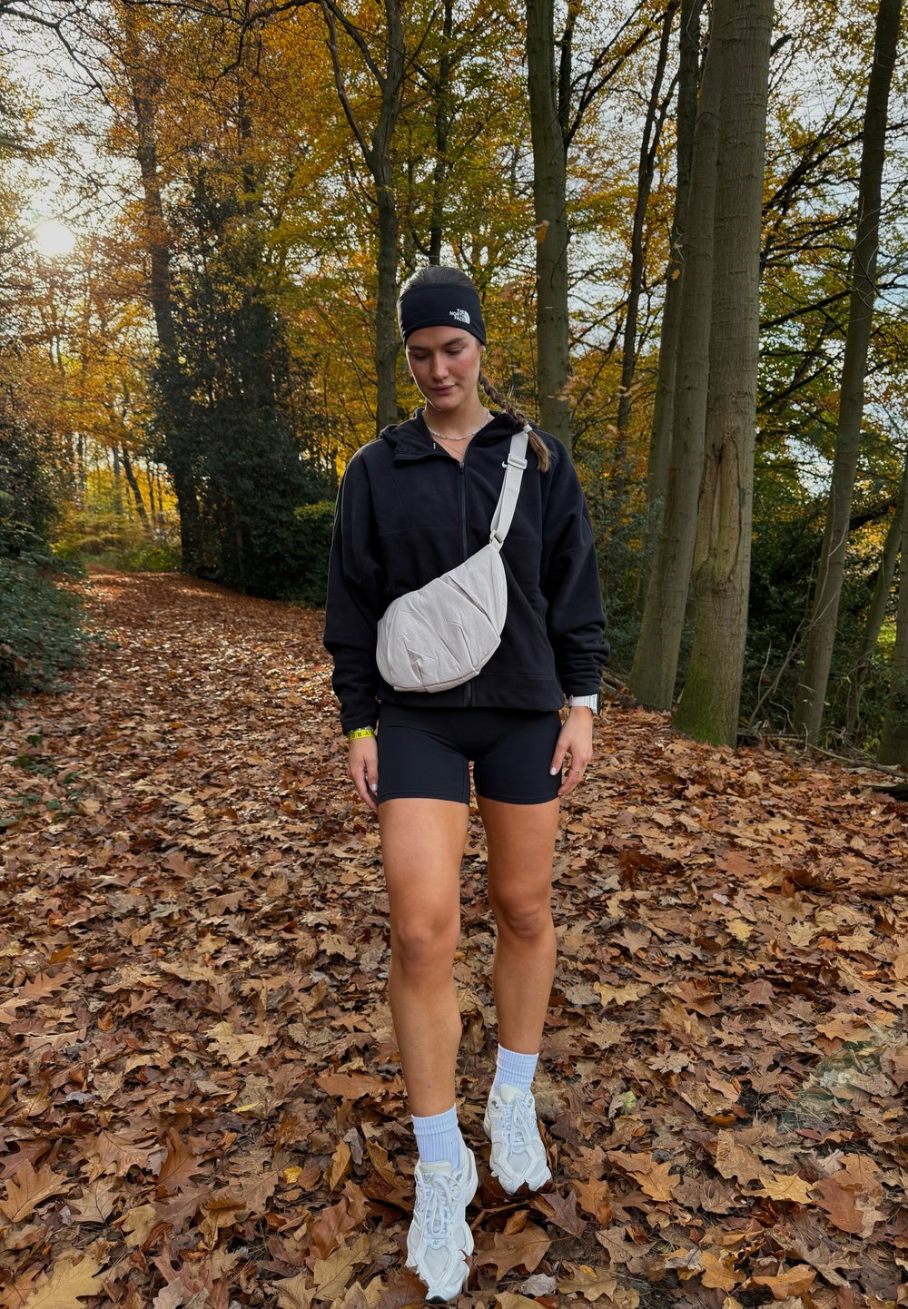 Lys grå nylon skulderveske båret over en svart glidelåsjakke, kombinert med svarte shorts og hvite joggesko, omgitt av høstløv.