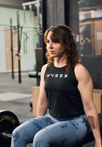 Haut de sport noir sans manches avec le texte blanc « HYROX », accompagné de leggings bleus marbrés. Tatouage notable de motif de pop-corn sur l'avant-bras.