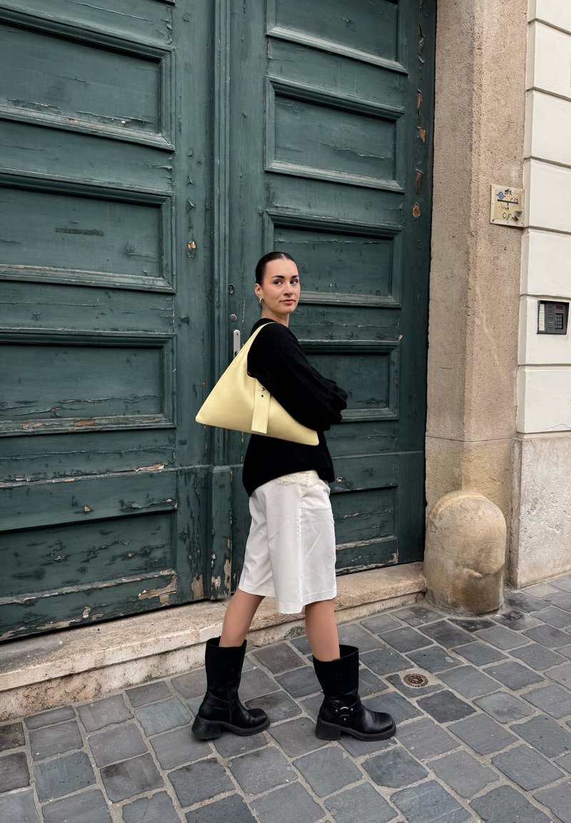 Black knitted sweater, white shorts, and black knee-high boots; carrying a yellow triangular bag; standing against a weathered green door.