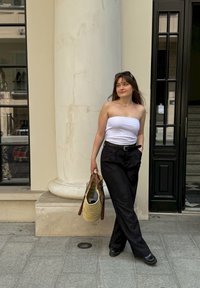 Μαύρο παντελόνι με φαρδιά πόδια και ψηλή μέση, συνδυασμένο με ένα λευκό μανίκι τοπ. Μια πλεκτή τσάντα από τερακότα με καφέ χερούλια κρατιέται σε ένα χέρι.