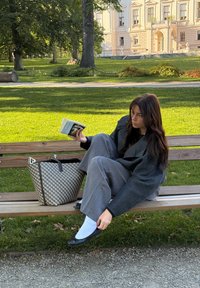 Žena u tamnom oversized kaputu i sivim hlačama sjedi na klupi, držeći knjigu. Pored nje se nalazi torba s uzorkom.