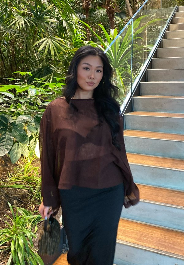 Kvinne med langt svart hår iført en brun gjennomsiktig bluse og svart skjørt, holder en liten veske, står ved inneplanter og trapp.