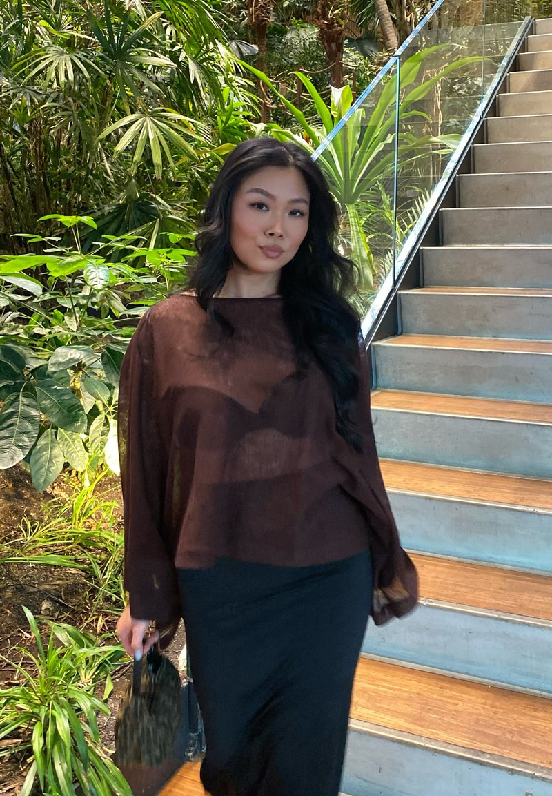 Femme aux longs cheveux noirs portant un chemisier marron transparent et une jupe noire, tenant un petit sac, debout près de plantes d'intérieur et d'un escalier.