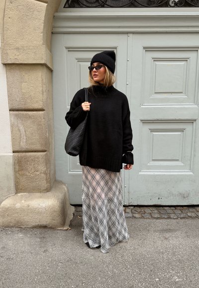 Suéter negro de gran tamaño con cuello alto, combinado con una falda larga y translúcida de cuadros en tonos blanco y gris. Los accesorios incluyen un gorro negro y gafas de sol.
