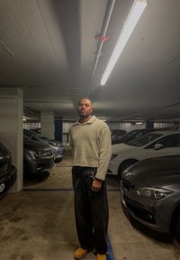 Homme en pull beige et pantalon noir, debout dans un parking souterrain faiblement éclairé, entouré de voitures garées.