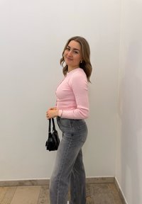 Rózsaszín bordázott hosszúujjú felső és szürke magas derekú farmernadrág, egy kis fekete táskával a kezében. Egy sima fehér fal előtt áll.