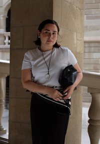 T-shirt bianco a maniche corte, gonna nera con un motivo a righe, borsa nera e una lunga collana d'argento. In piedi contro una colonna di pietra.