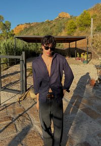 Un hombre con una camisa de lino azul oscuro y jeans negros está de pie en un camino de piedra. Lleva gafas de sol y una cadena de plata, con un estilo minimalista.