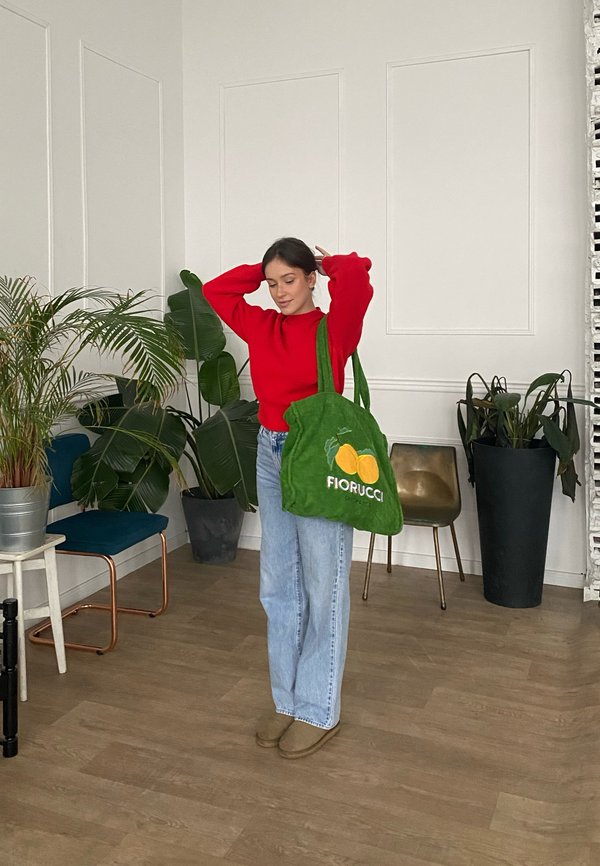 Bolso tote verde con el logo "Fiorucci" y un gráfico de limón, llevado al hombro. Modelo con suéter rojo y jeans azules, en un entorno interior con plantas.