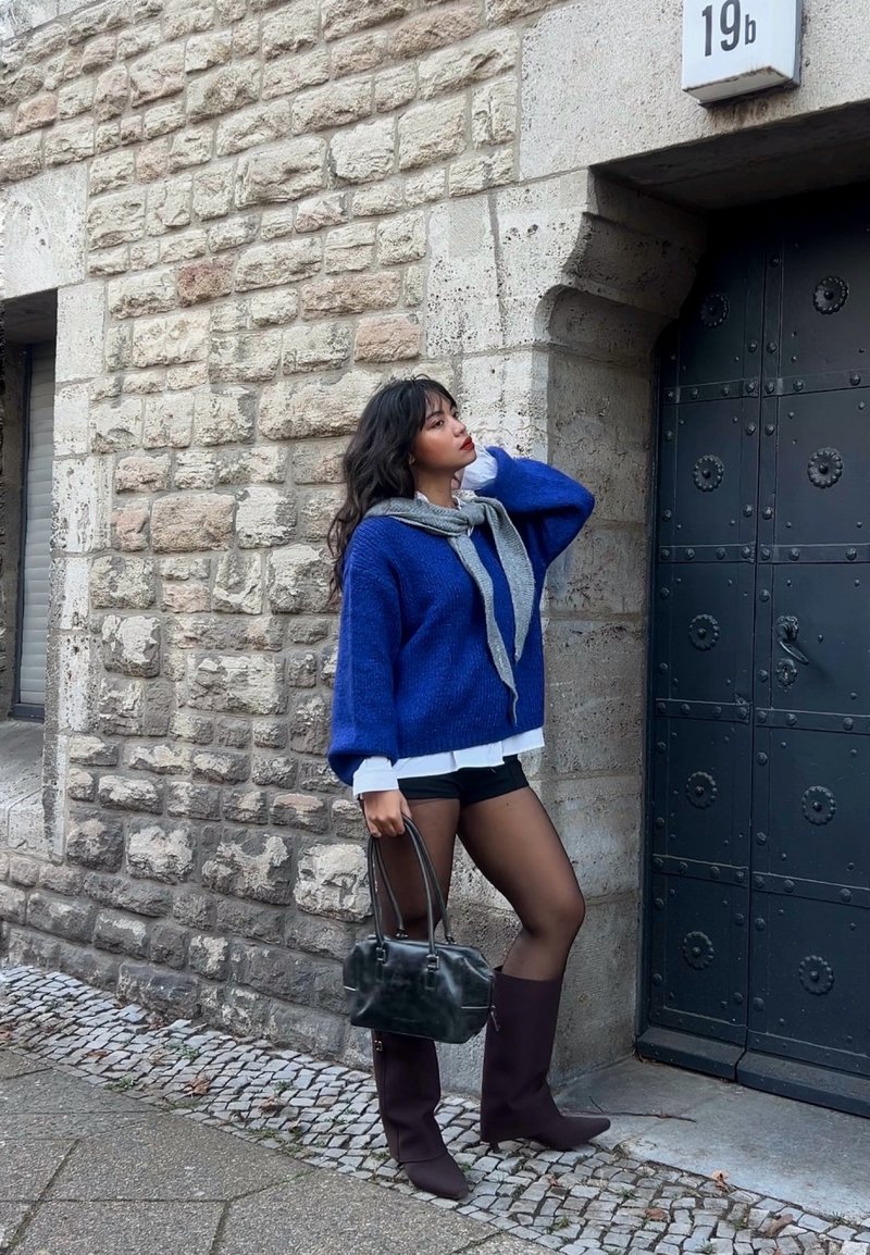 Maglione blu oversize, sciarpa grigia annodata al collo, pantaloncini neri, borsa nera, stivali marroni al ginocchio, sfondo con muro di pietra strutturata.