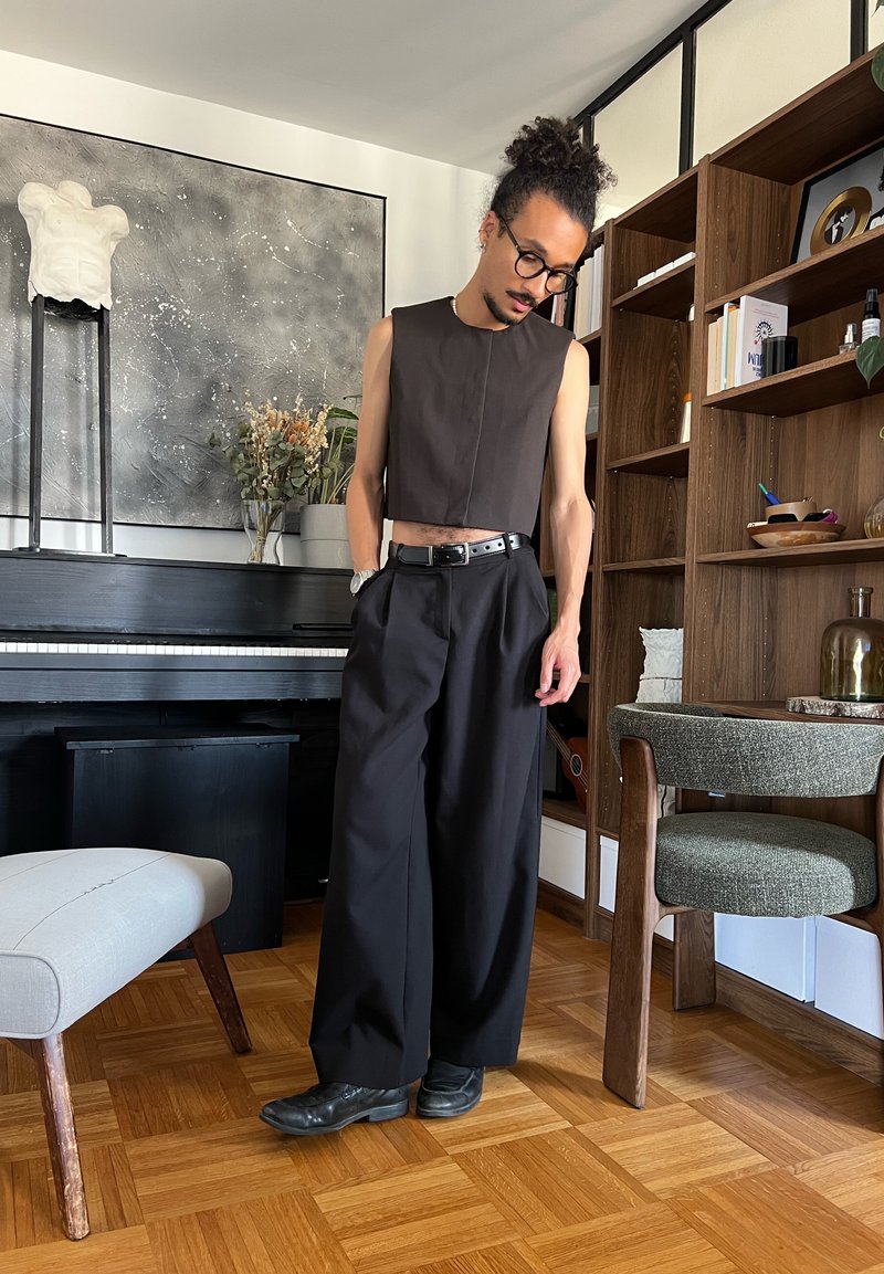 Homme aux cheveux bouclés attachés, portant des lunettes, un haut noir sans manches et un pantalon noir large, debout dans un salon avec un piano et une bibliothèque.