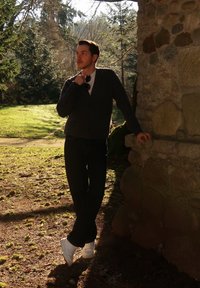 Jeune homme en vêtements sombres et baskets blanches appuyé contre un mur de pierre en plein air, tenant des lunettes de soleil, avec des arbres et de l'herbe au soleil derrière lui.