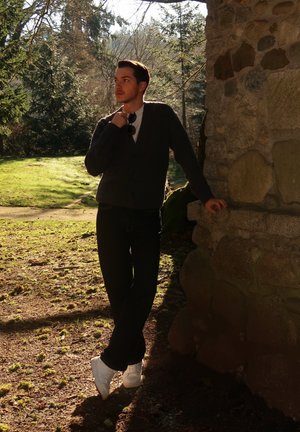 Joven con ropa oscura y zapatillas blancas apoyado contra una pared de piedra al aire libre, sosteniendo gafas de sol, con árboles y césped iluminados por el sol detrás de él.