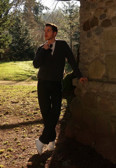 Joven con ropa oscura y zapatillas blancas apoyado contra una pared de piedra al aire libre, sosteniendo gafas de sol, con árboles y césped iluminados por el sol detrás de él.