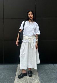 Witte katoenen t-shirt, geknoopt in de taille met een witte sjerp, gecombineerd met een vloeiende witte rok en zwarte houten klompen; zwarte schoudertas.