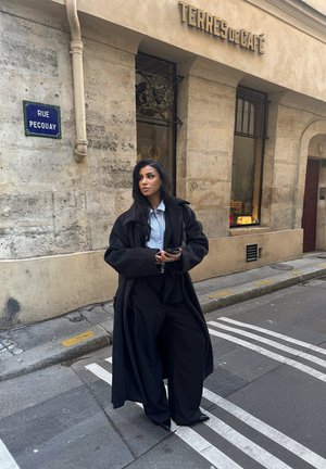 Abrigo negro largo sobre una camisa azul clara, combinado con pantalones anchos negros. El fondo presenta un edificio de piedra y una señal de tráfico.