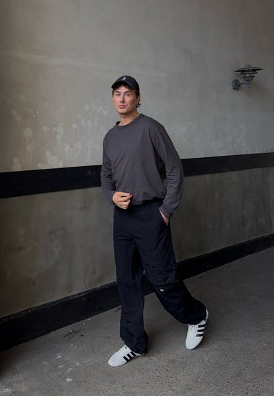 Camisa de manga larga gris y pantalones cargo negros con bolsillos. Zapatillas blancas con rayas negras. Fondo simple con paredes texturizadas.