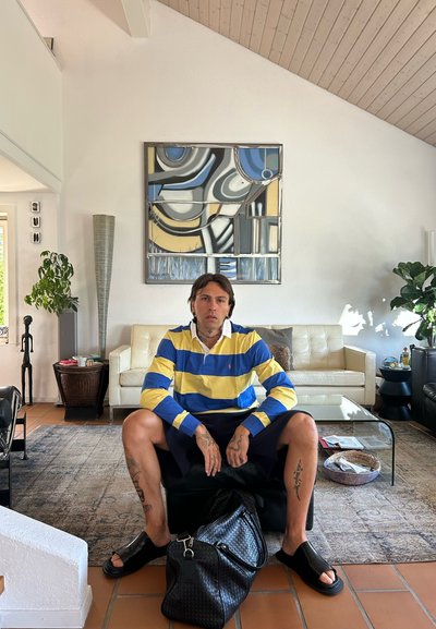 Camisa estilo rugby con rayas azules y amarillas, pantalones cortos negros y sandalias negras. Sentado sobre una bolsa negra en un ambiente moderno de sala de estar.