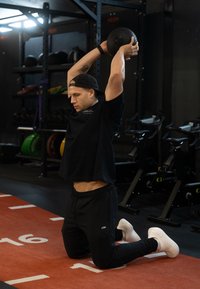 Atlét v čiernom tričku a nohaviciach vykonáva cvičenie s kettlebellom, kľačiac na oranžovej podložke v telocvični s vybavením v pozadí.