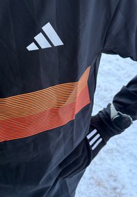 Personne portant une veste noire Adidas avec des rayures orange, des gants noirs avec des rayures blanches, et une montre connectée blanche, debout sur la neige.