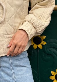 Beige gesteppte Jacke mit strukturiertem Finish, elastischen Bündchen und Kordelzug. Getragen über hellblauer Jeans. Sonnenblumenmotiv im Hintergrund.