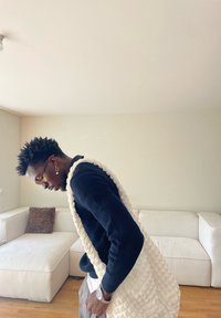 Mężczyzna w czarnym swetrze, z okularami i kolczykiem, noszący dużą, teksturowaną białą torbę na ramię w jasnym salonie z białą kanapą.