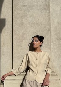 Beige linneknappetopp med rund hals og trekvartlange ermer, med en buet skjortehem og naturlige trematerialer.