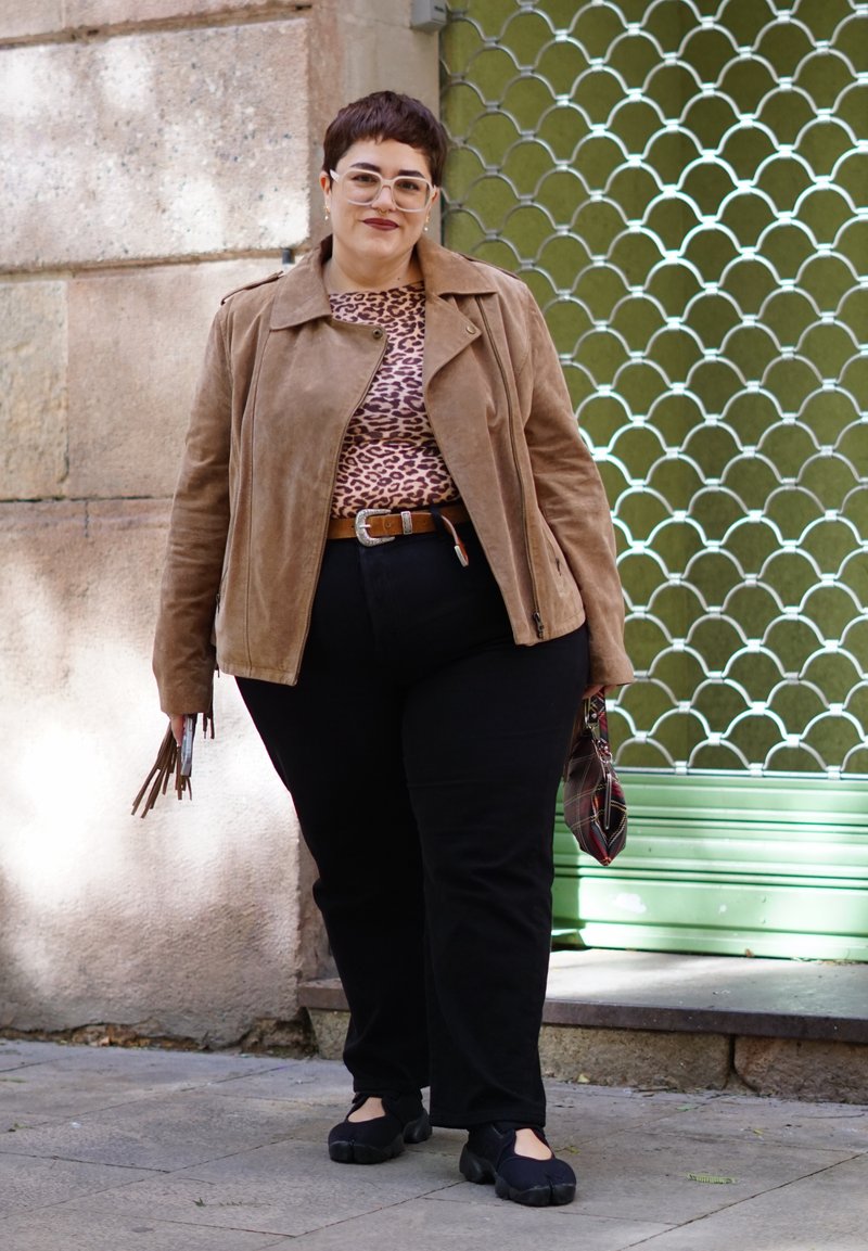 Personne aux cheveux courts portant des lunettes transparentes, une veste marron, un haut à imprimé léopard, un pantalon noir, tenant un sac et marchant sur le trottoir.