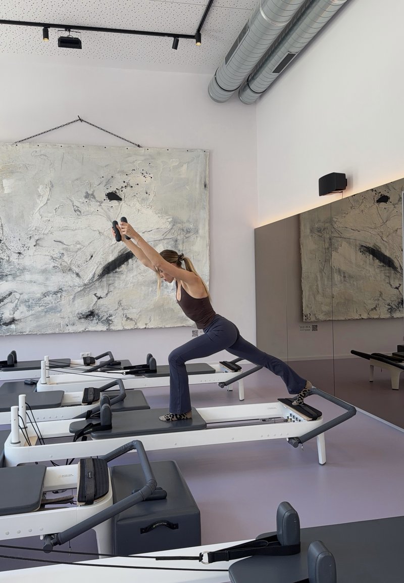 Eine Frau in Sportkleidung dehnt sich nach vorne auf einem Pilates-Reformer in einem hellen Studio mit abstrakter Kunst und Spiegeln.